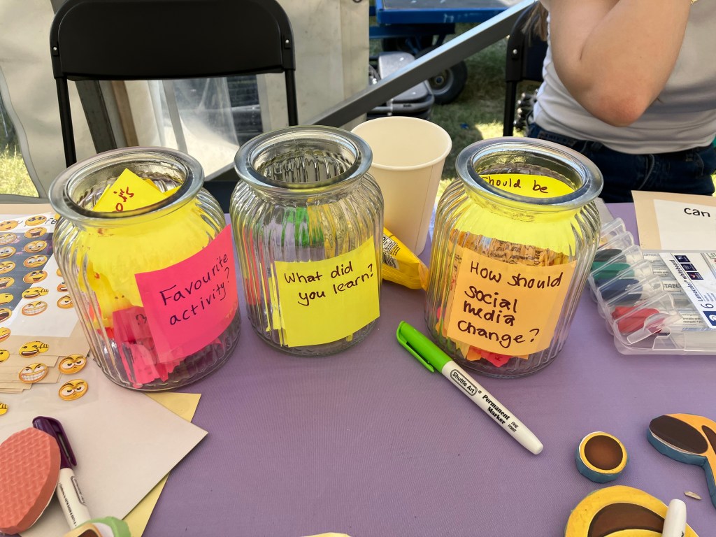 Three transparent jars for collecting participant feedback notes. The first jar from the right is titled ‘Favourite activity?’, the middle one ‘What did you learn?’ and the third one ‘How should social media change?’